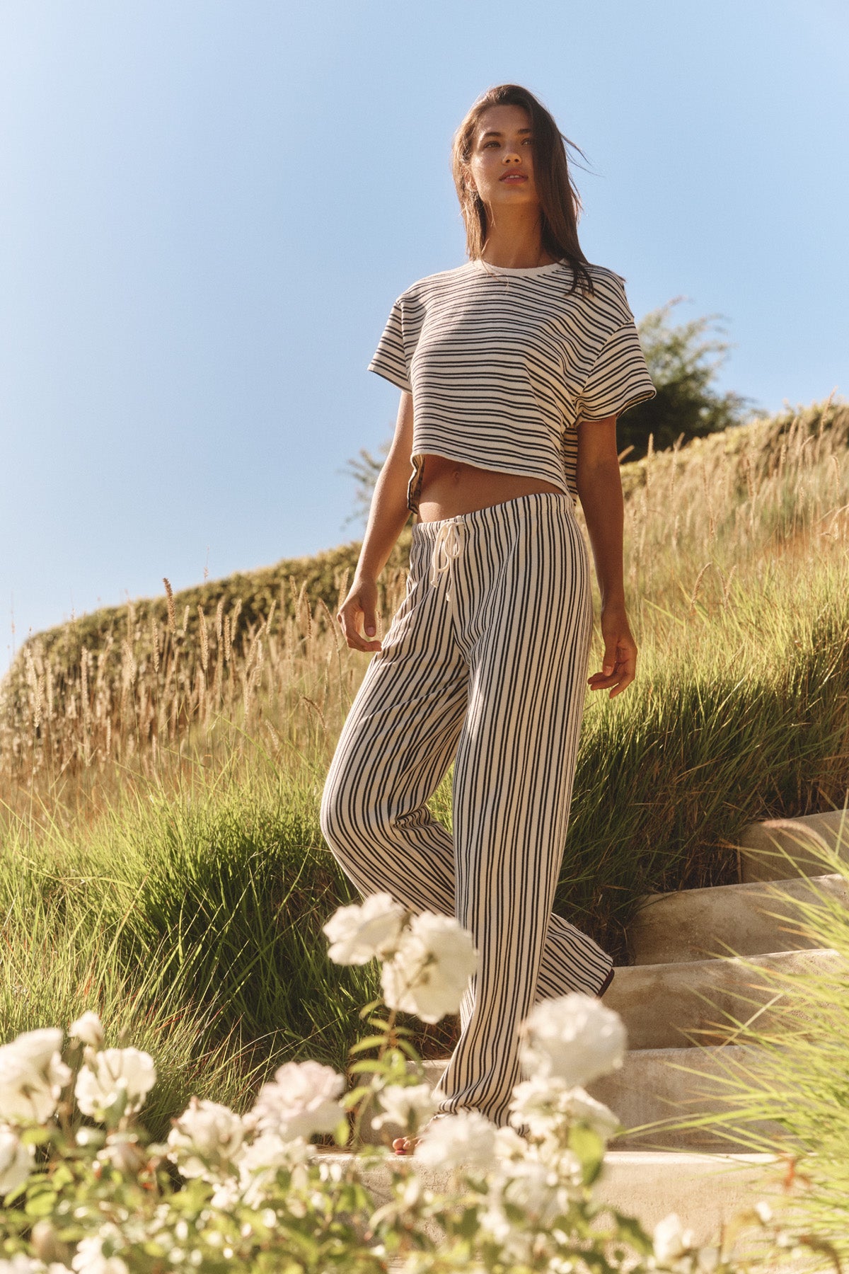 A woman in Velvet by Graham & Spencer’s AMELISE SWEATPANT with a matching striped crop top stands on outdoor steps amid grass and white flowers under a clear sky.-40909663174849
