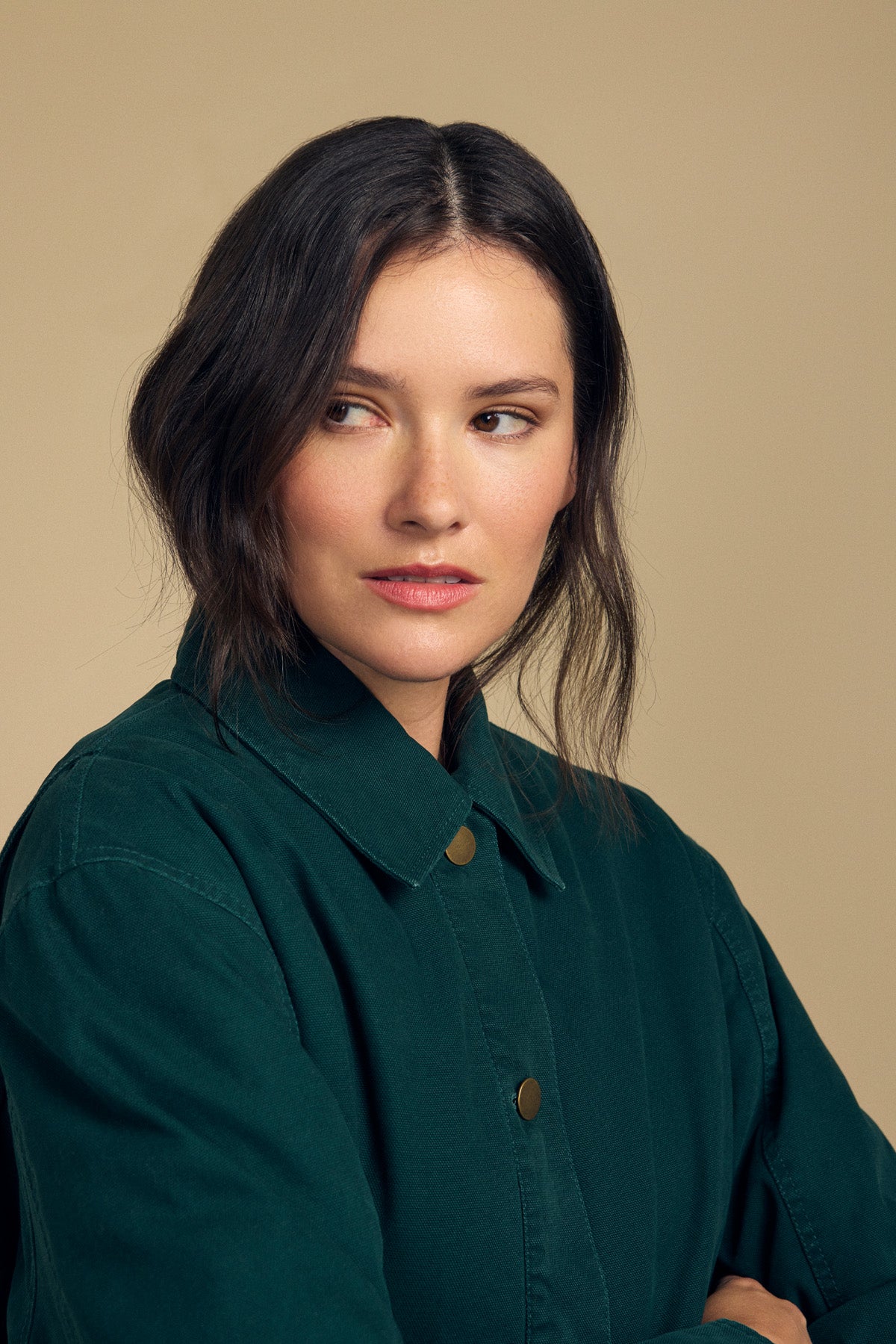 A woman with shoulder-length dark hair wears the Velvet by Graham & Spencer DYLAN BARN JACKET in dark green, gazing slightly to the side against a beige background.-41815026532545