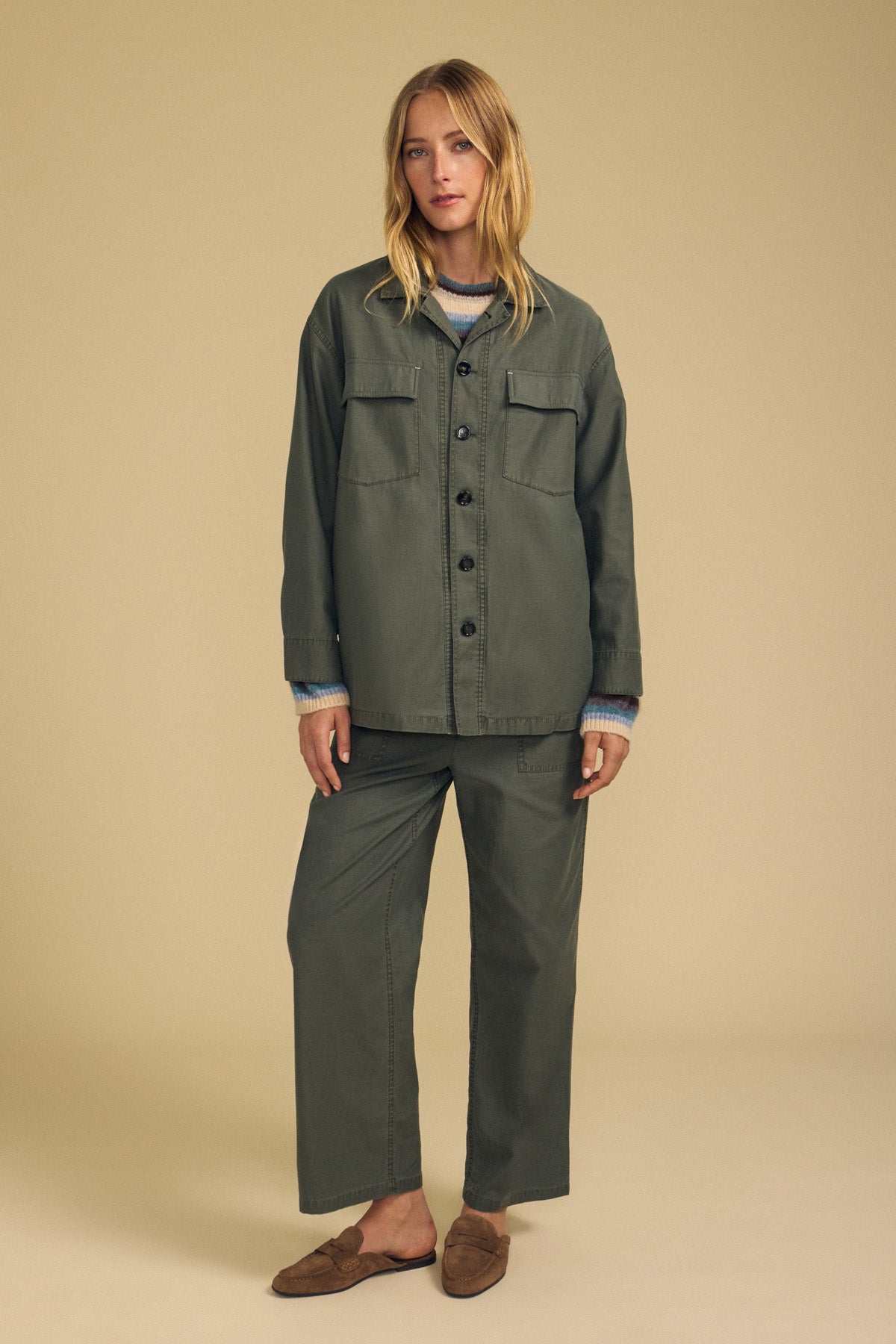 A woman stands facing forward in a beige studio, wearing the KAY SHIRT JACKET by Velvet by Graham & Spencer in olive green brushed cotton, paired with matching wide-leg pants, layered over a blue and white sweater, and finished with brown slip-on shoes.-41964575817921