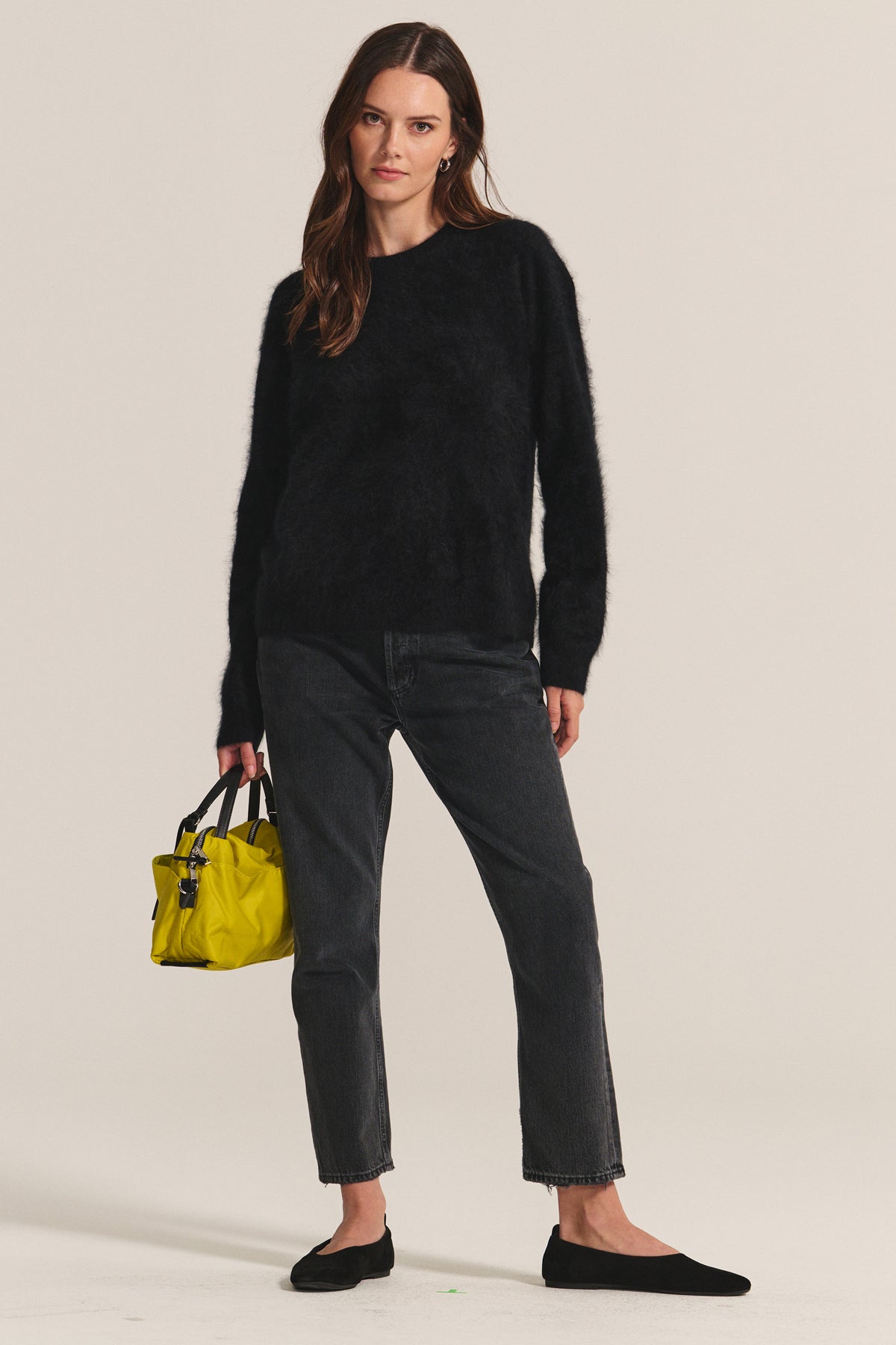 A woman stands against a plain background wearing the JACQUELINE CASHMERE SWEATER by Velvet by Graham & Spencer, black jeans, black flats, and holding a small yellow handbag.-40247688495297