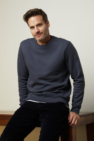 A man sitting on a sturdy wooden table wearing a clean Velvet by Graham & Spencer KING CREW NECK SWEATSHIRT.