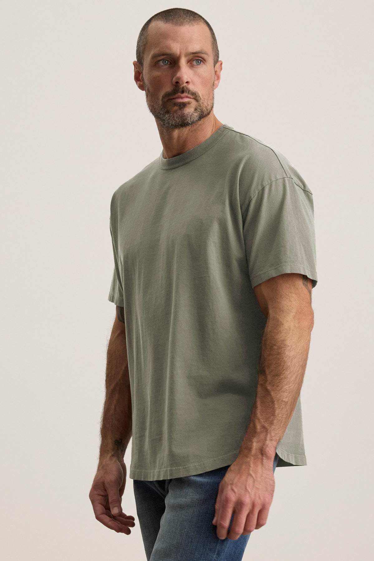 A man poses against a neutral backdrop in the relaxed-fit BRYAN TEE by Velvet by Graham & Spencer, paired with blue jeans, showcasing an ensemble that effortlessly blends style and comfort for everyday versatility.