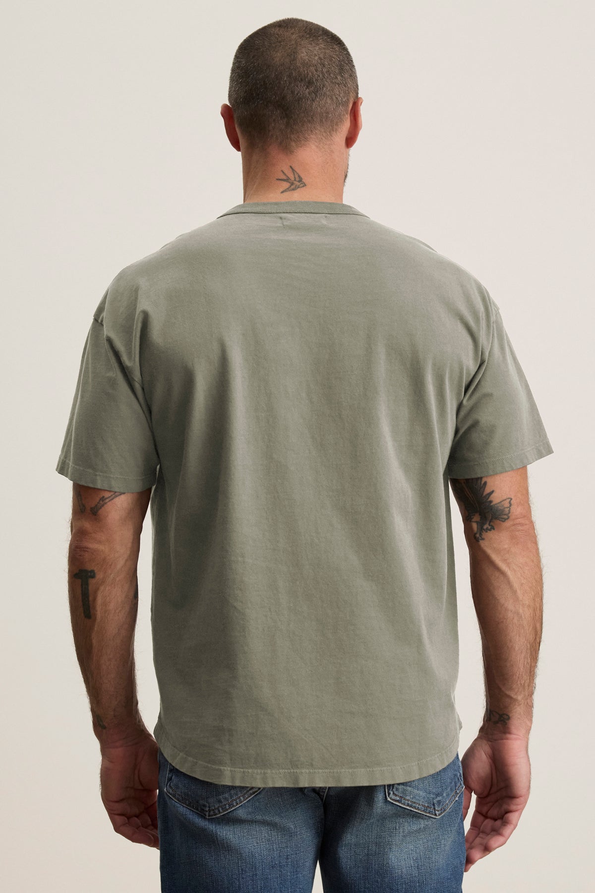 A man in a relaxed fit BRYAN TEE by Velvet by Graham & Spencer and jeans, viewed from behind. His shaved head and tattoos on his arms and neck effortlessly reflect everyday versatility.