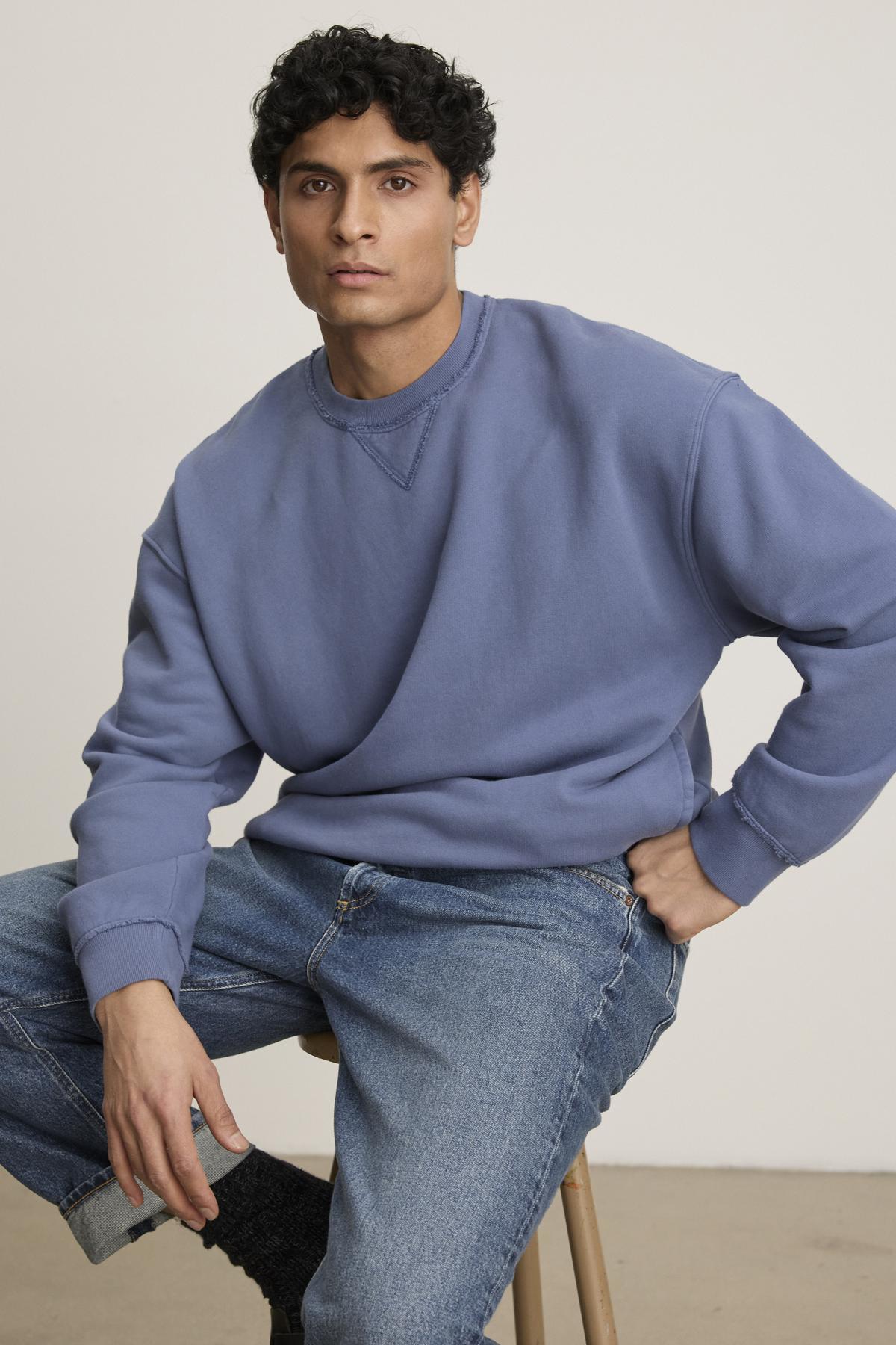 A person lounges in Velvet by Graham & Spencer's FORREST SWEATSHIRT—a blue heavyweight cotton fleece with ribbed trims and relaxed fit—paired with jeans while sitting on a stool against a plain backdrop, exuding casual comfort.
