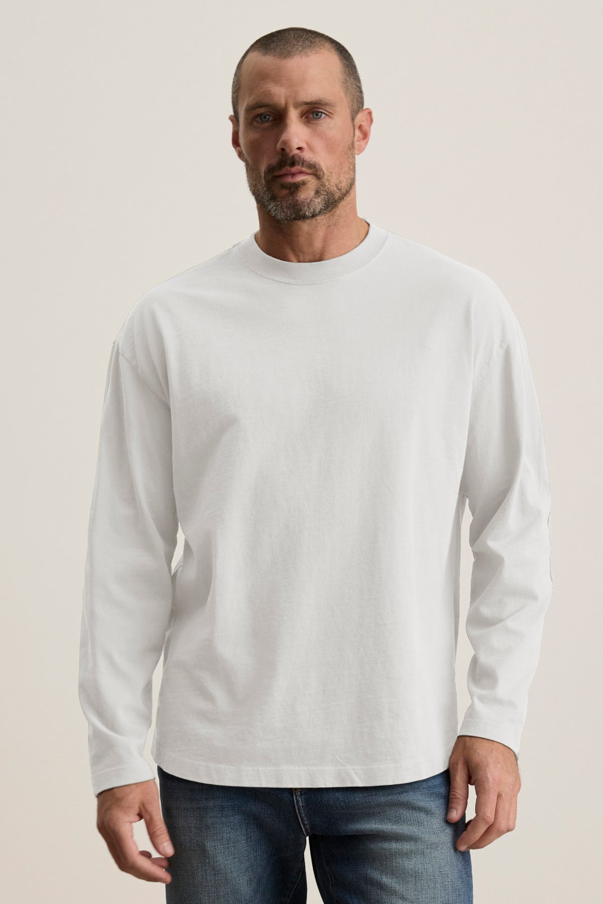 A man with a short beard wears the BRYAN TEE, a plain white long-sleeve shirt in structured pima cotton from Velvet by Graham & Spencer, paired with relaxed fit blue jeans. He stands against a neutral background, showcasing everyday versatility.-39018195321025