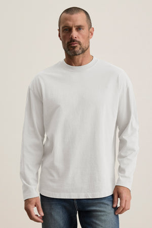 A man with a short beard wears the BRYAN TEE, a plain white long-sleeve shirt in structured pima cotton from Velvet by Graham & Spencer, paired with relaxed fit blue jeans. He stands against a neutral background, showcasing everyday versatility.