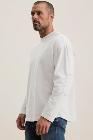 A man with short hair and a beard wears a plain white long-sleeve BRYAN TEE from Velvet by Graham & Spencer in structured pima cotton and blue jeans, highlighting everyday versatility against a neutral background.