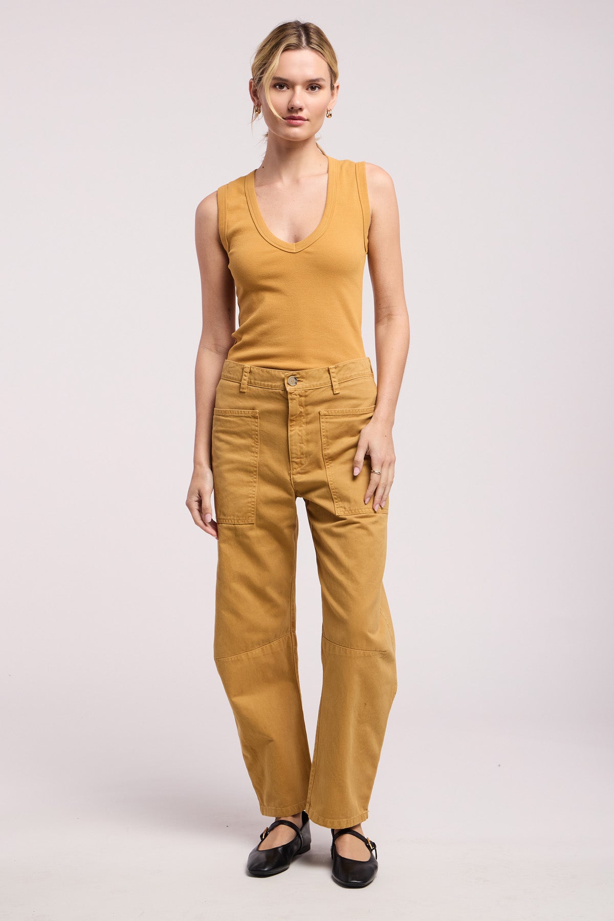 A woman stands against a plain background, wearing a sleeveless tan top, the BRYLIE PANT by Velvet by Graham & Spencer with utilitarian patch pockets, and black flat shoes.-41374540398785