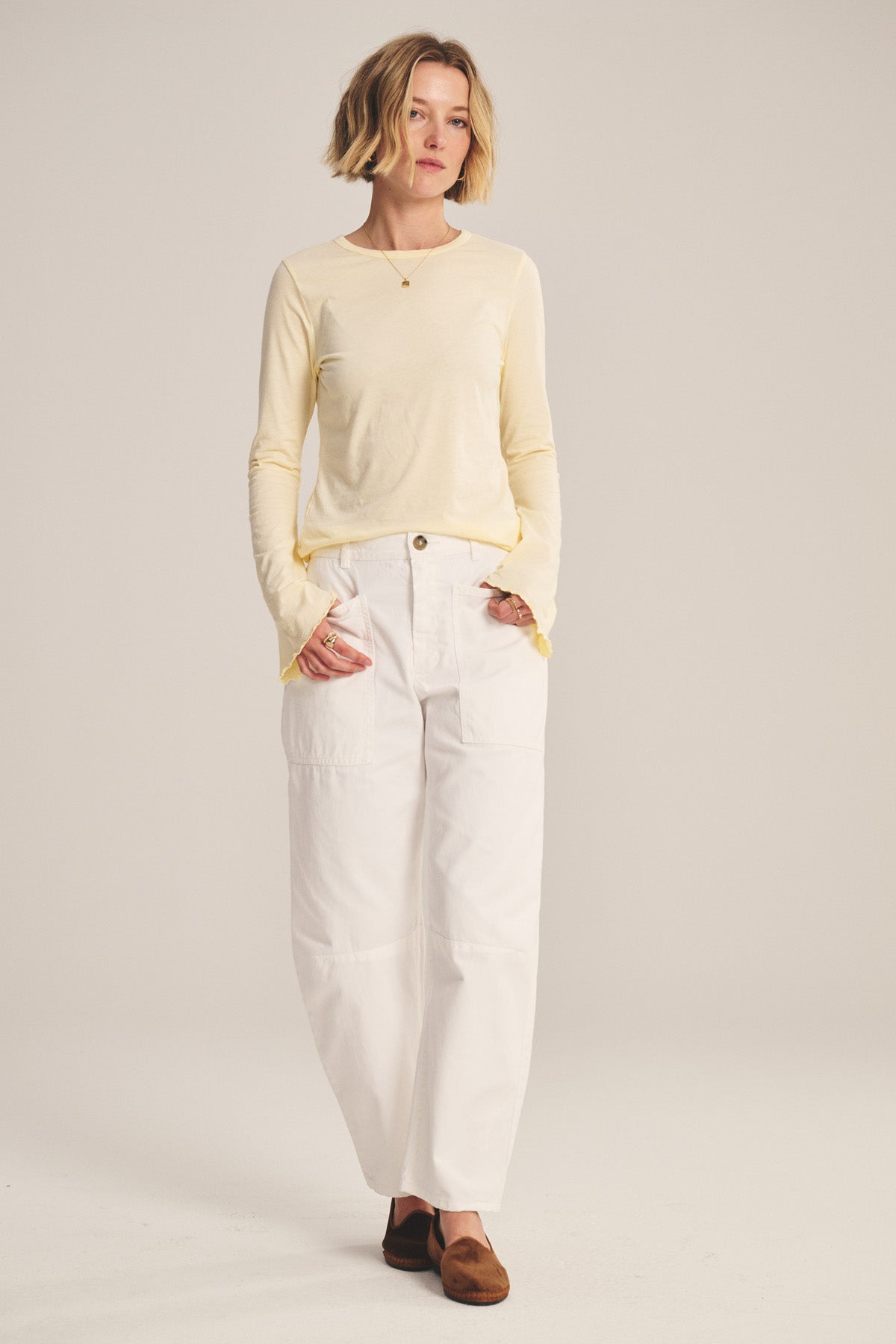 A woman stands with hands in pockets, wearing the BRYLIE PANT by Velvet by Graham & Spencer—white cotton twill trousers with utilitarian patch pockets—paired with a light yellow long-sleeve shirt and brown shoes against a plain backdrop.-40959929123009