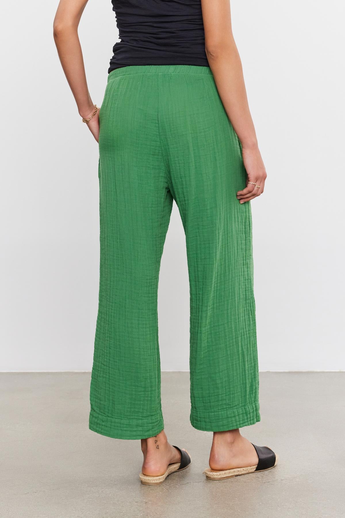 A person standing with their back to the camera, wearing green FRANNY COTTON GAUZE PANTS by Velvet by Graham & Spencer and black flat shoes.