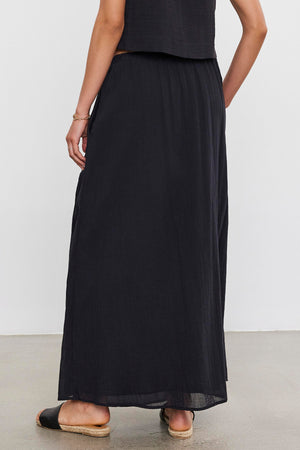 Woman in a black INDY COTTON GAUZE SKIRT and flat shoes, viewed from the back, standing against a neutral background.