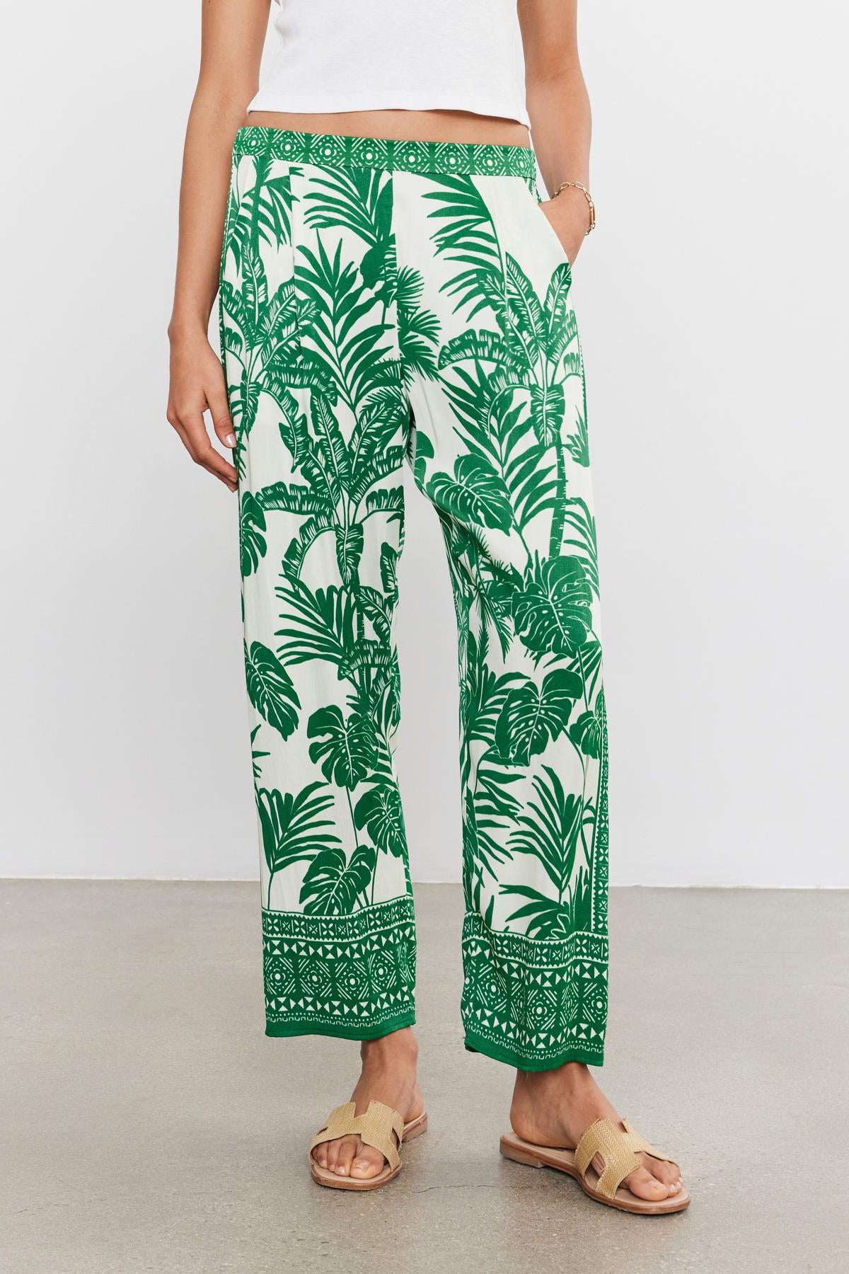Woman standing in Velvet by Graham & Spencer's IRIS PANT in white and green palm print with an elastic waist, paired with a plain white top and beige sandals, against a neutral background.-36917074100417