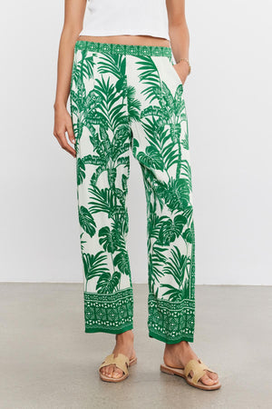 Woman standing in Velvet by Graham & Spencer's IRIS PANT in white and green palm print with an elastic waist, paired with a plain white top and beige sandals, against a neutral background.
