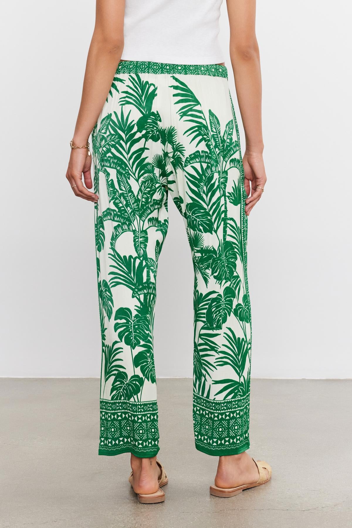 A person wearing Velvet by Graham & Spencer's IRIS PANT with a patterned green and white palm print pants with a tropical leaf design, viewed from the back.-36917074198721