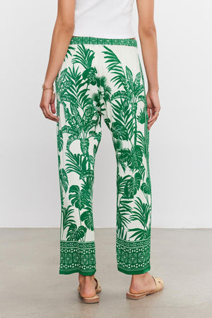 A person wearing Velvet by Graham & Spencer's IRIS PANT with a patterned green and white palm print pants with a tropical leaf design, viewed from the back.