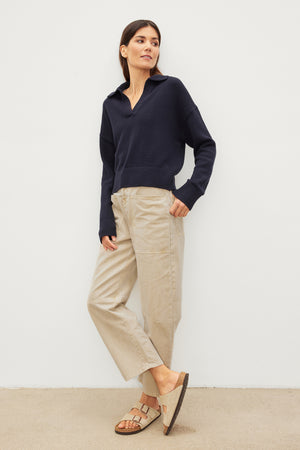 Woman in a navy blue sweater and beige Velvet by Graham & Spencer BRYLIE SANDED TWILL UTILITY PANT leaning against a white wall.