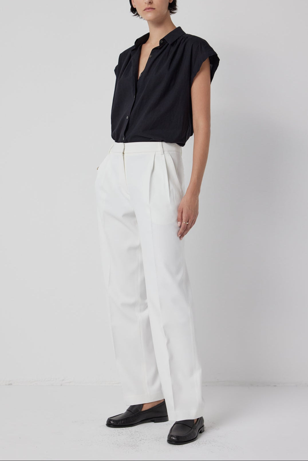 A woman standing against a white backdrop, wearing a black blouse, white Velvet by Jenny Graham relaxed fit BUNDY PANT trousers, and black loafers.-36524163563713
