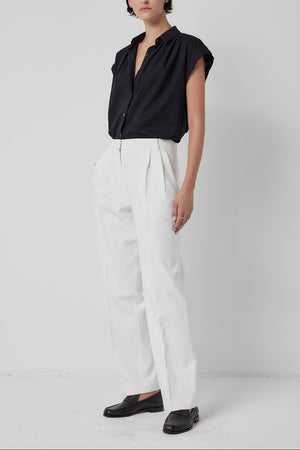 A woman standing against a white backdrop, wearing a black blouse, white Velvet by Jenny Graham relaxed fit BUNDY PANT trousers, and black loafers.