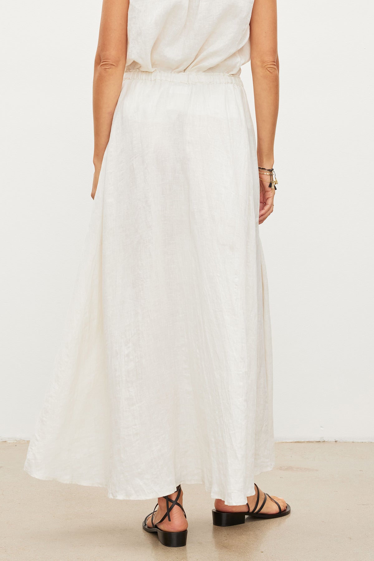 A person standing with their back facing the camera, wearing the BAILEY LINEN MAXI SKIRT by Velvet by Graham & Spencer and black sandals, showcasing an elegant woven linen fabric.