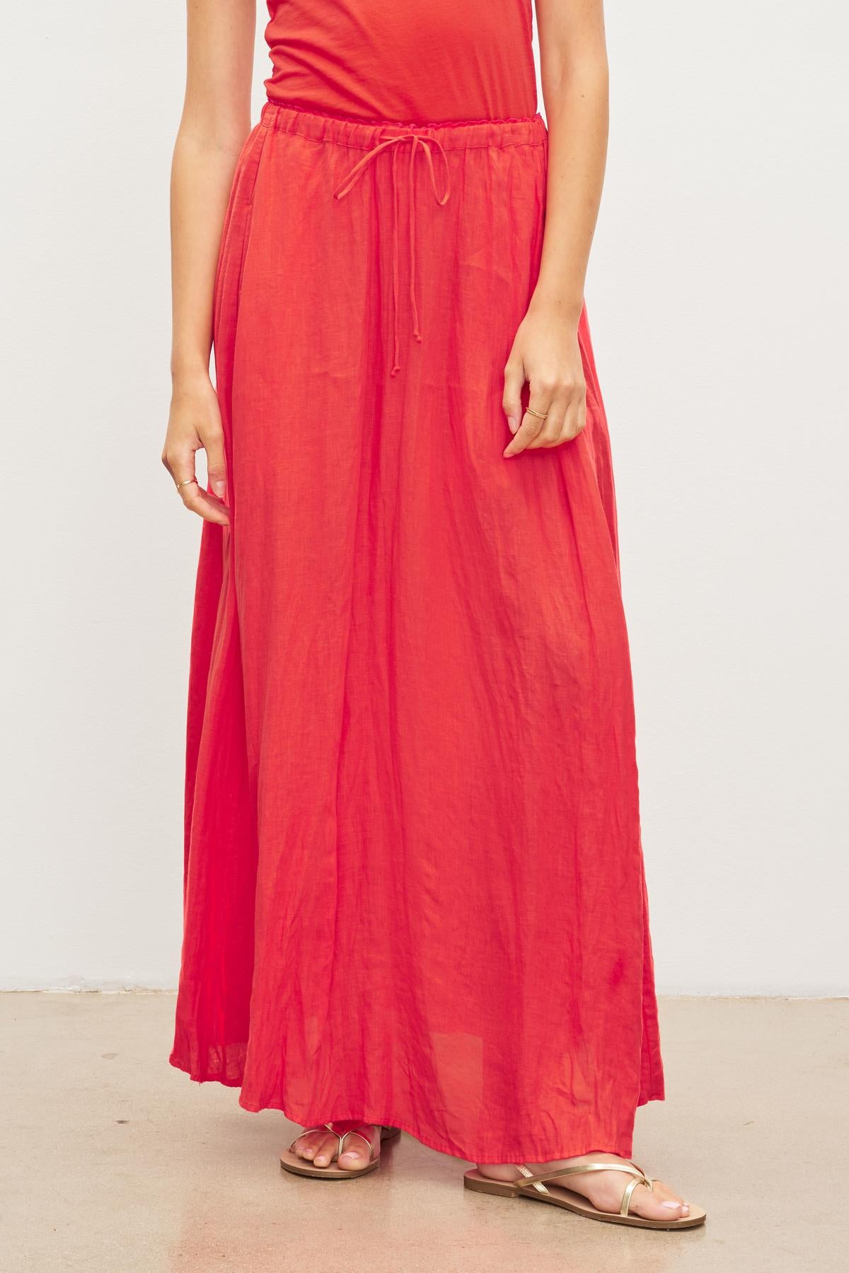 Wearing the Velvet by Graham & Spencer BAILEY LINEN MAXI SKIRT, a person in a red linen skirt with an elastic drawstring waist and gold sandals stands against a plain white background.