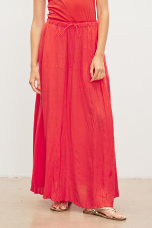 Wearing the Velvet by Graham & Spencer BAILEY LINEN MAXI SKIRT, a person in a red linen skirt with an elastic drawstring waist and gold sandals stands against a plain white background.