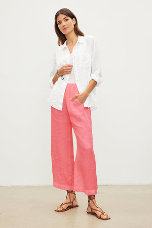 A woman stands against a plain white background, wearing a white button-up shirt and pink Velvet by Graham & Spencer LOLA LINEN PANT with an elastic waist. She has brown hair and is wearing sandals.