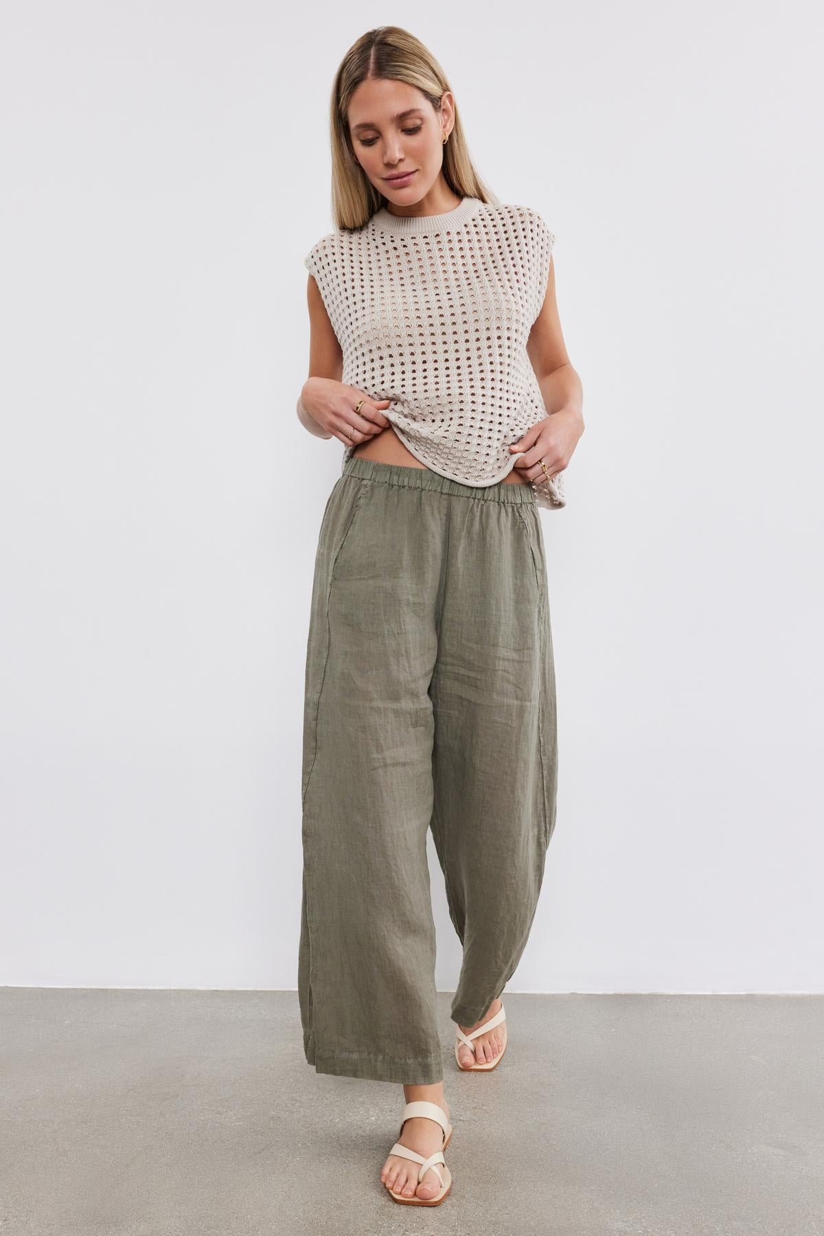 A woman stands against a plain background, tucking a Velvet by Graham & Spencer MAISON SWEATER into loose green trousers, looking down, with her hair styled down.-36910015905985