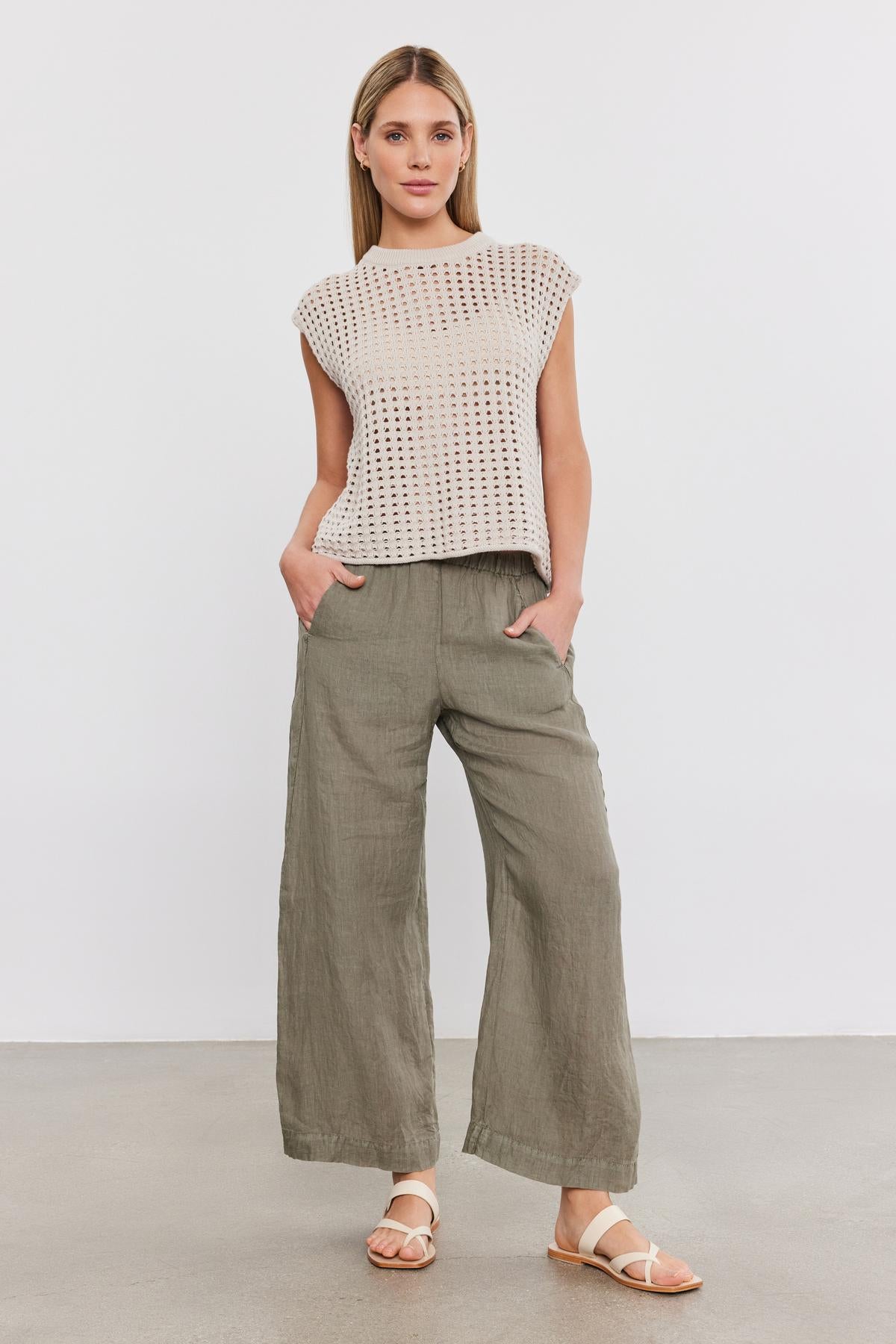 A woman stands in a studio, wearing a white cotton cashmere mesh knit top by Velvet by Graham & Spencer and gray wide-leg trousers with white sandals.-36910015938753