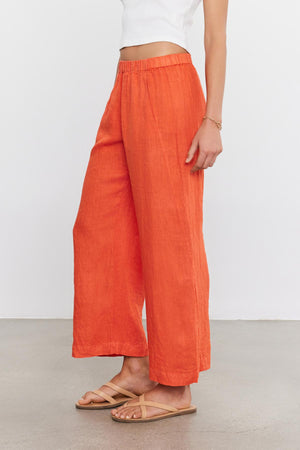 A person is wearing Velvet by Graham & Spencer LOLA LINEN PANT, a white crop top, brown sandals, and a bracelet, standing sideways against a plain background.