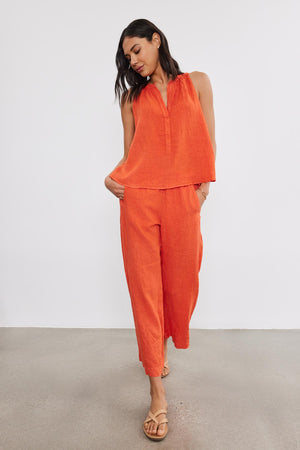 A person is standing indoors wearing an orange sleeveless top and matching relaxed leg pants. They have their hands in their pockets and are looking down. Their footwear is light brown sandals.