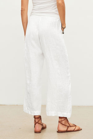 A person stands on a beige floor, seen from the back, wearing a white sleeveless top and Velvet by Graham & Spencer's LOLA LINEN PANT—lightweight linen pull-on trousers with relaxed legs—paired with brown strappy sandals.