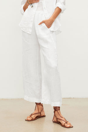 Model wearing the LOLA LINEN PANT by Velvet by Graham & Spencer, paired with a white button-up shirt and brown strappy sandals, standing against a plain light background.