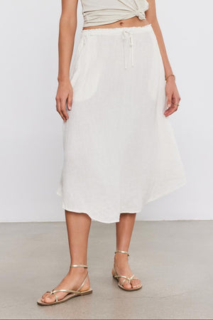 A woman stands in a studio wearing a beige wrap top and a NEMY LINEN SKIRT by Velvet by Graham & Spencer with an elastic waist, paired with gold sandals.