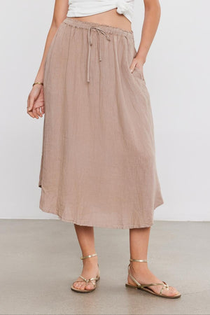 A woman in a white tank top and NEMY LINEN SKIRT by Velvet by Graham & Spencer stands against a plain background, wearing flat sandals.