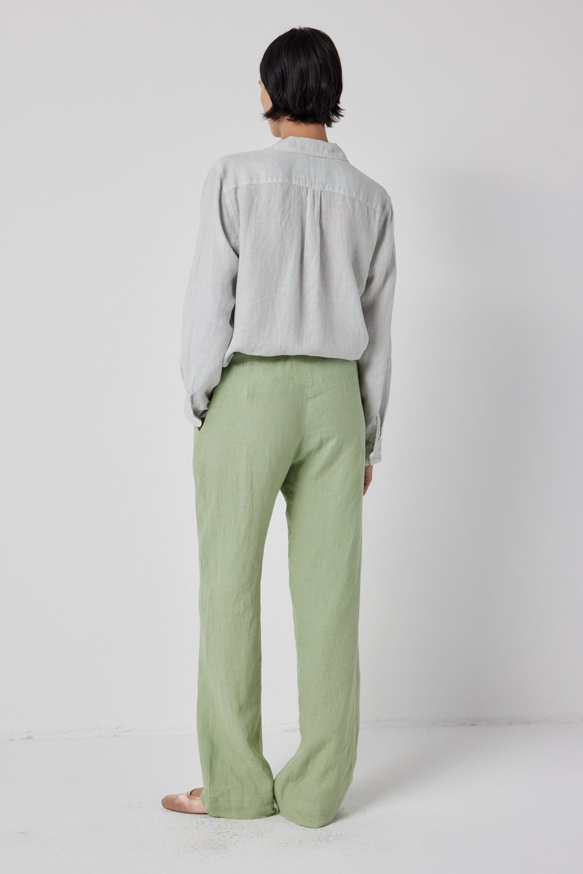   A person viewed from behind, wearing a loose gray blouse and Velvet by Jenny Graham's Pico Linen Pant in light green, standing against a plain white background. 