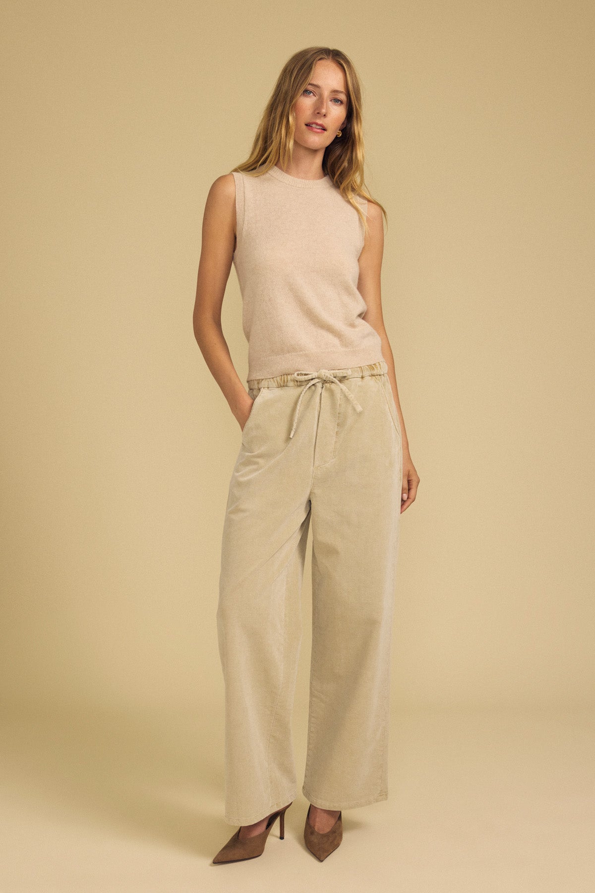 A woman poses against a beige background with her hands in her pockets and a relaxed expression, wearing the IVANA CASHMERE SWEATER VEST by Velvet by Graham & Spencer, paired with wide-leg beige drawstring pants and brown heels.-41964532334785