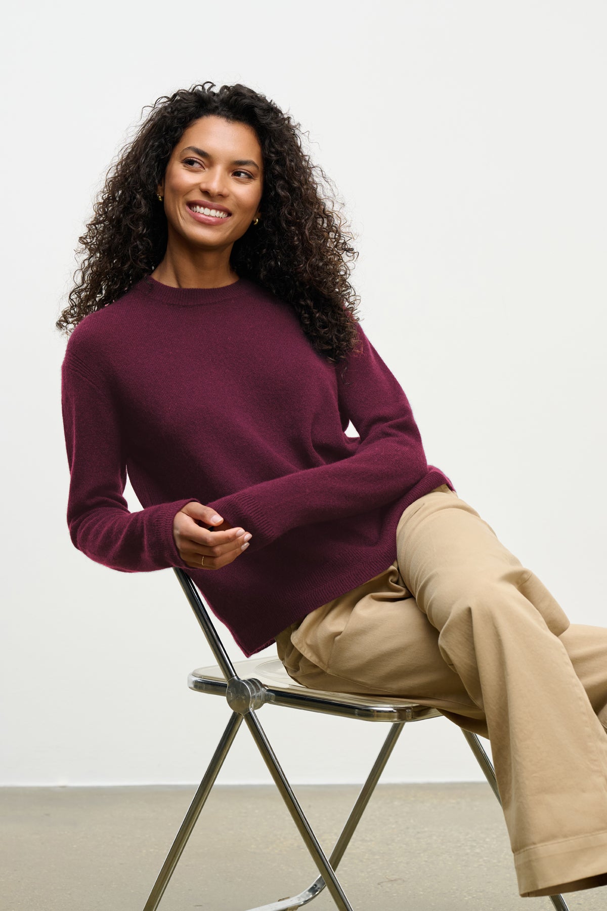A woman with curly hair wears the CRYSTAL CASHMERE SWEATER by Velvet by Graham & Spencer in maroon and beige pants, sitting on a metal chair against a plain light background, smiling slightly to the side.