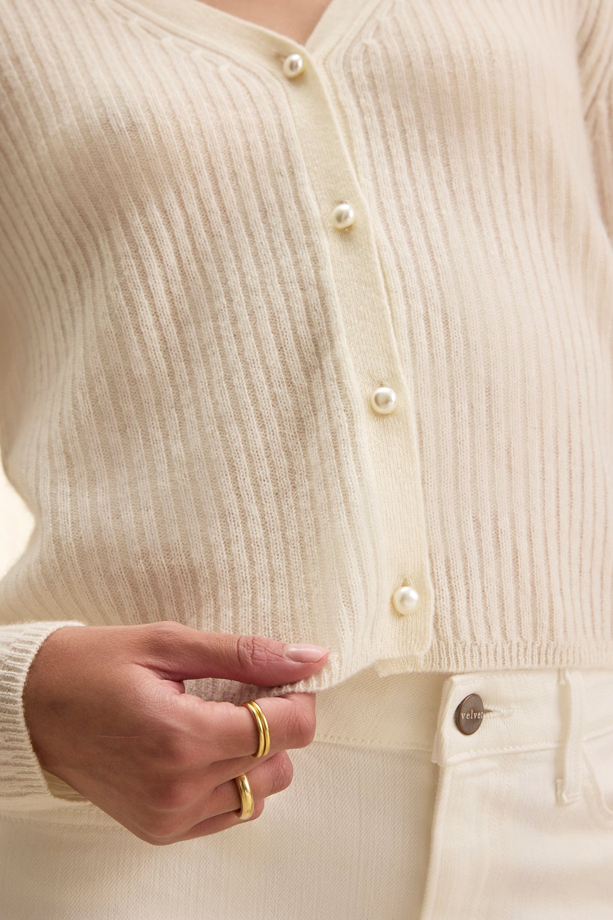 A person wearing the WES CASHMERE CARDIGAN by Velvet by Graham & Spencer, an off-white ribbed button-up with matching pants and gold rings, holding the edge of their cardigan.-39333168873665