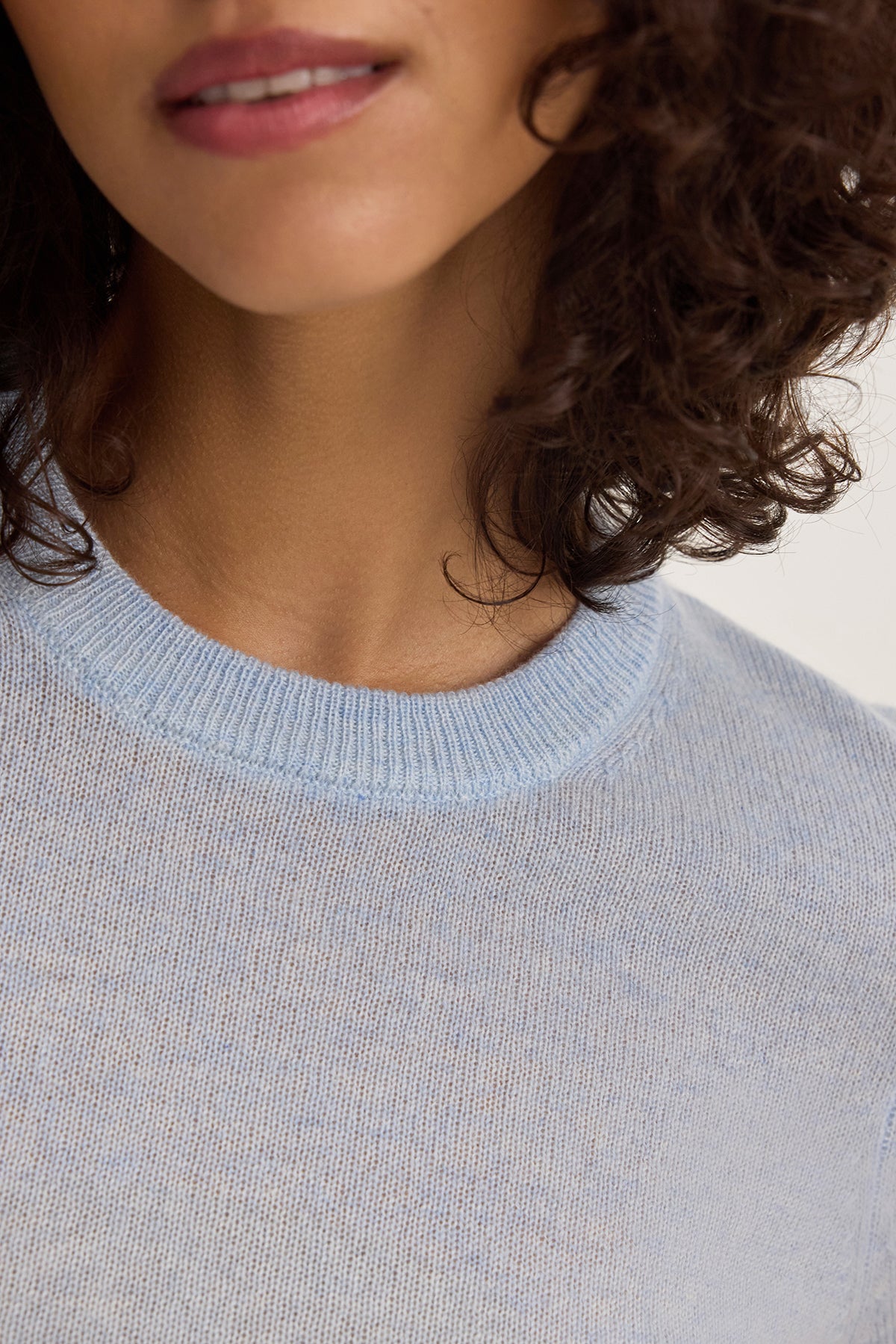 A person with curly hair is wearing the YARA CASHMERE SWEATER by Velvet by Graham & Spencer in light blue, featuring ribbed trims.-38663174095041