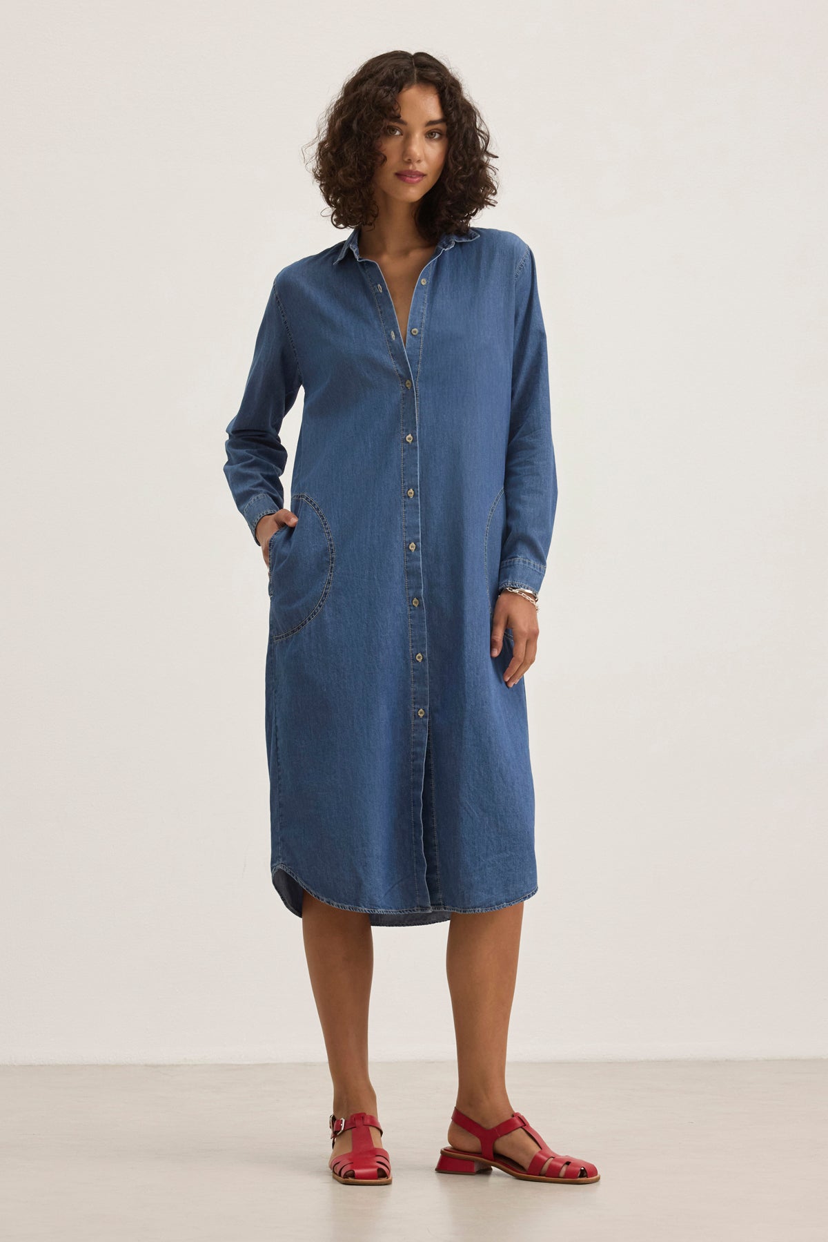 A person stands against a plain background wearing the IZMIR CHAMBRAY DRESS, a long-sleeve button-up denim dress by Velvet by Graham & Spencer, paired with red sandals.