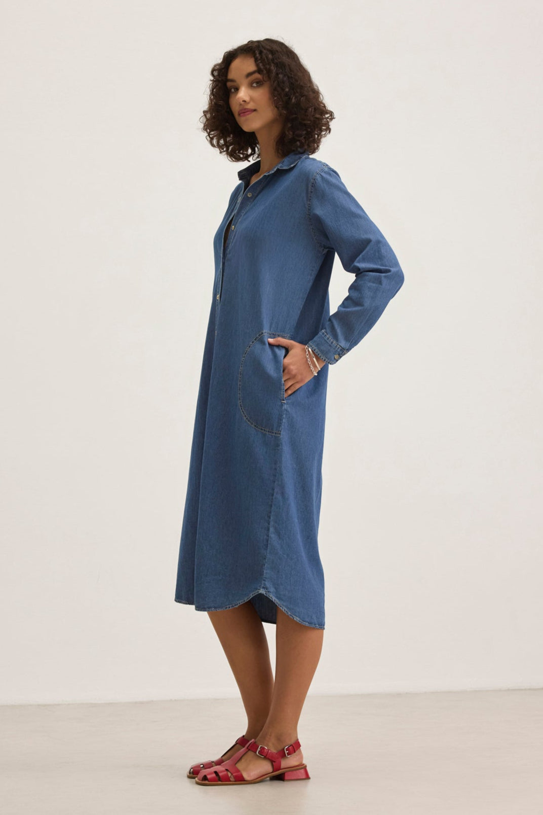 A person models the IZMIR CHAMBRAY DRESS from Velvet by Graham & Spencer, a blue denim long-sleeve dress with large pockets, paired with red sandals, set against a plain white backdrop.-38820646486209