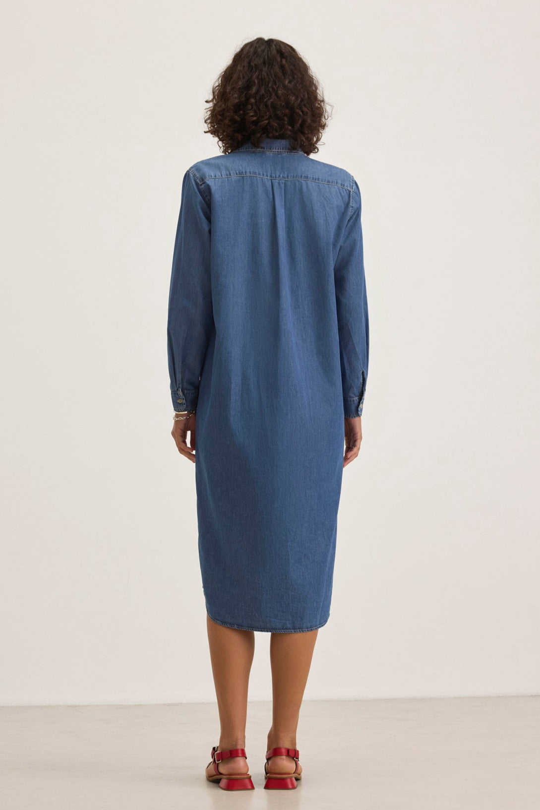 Back view of a person with curly hair wearing the IZMIR CHAMBRAY DRESS by Velvet by Graham & Spencer and red sandals, standing on a neutral background.-38820646420673