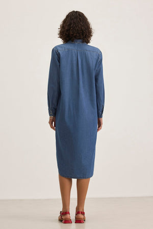 Back view of a person with curly hair wearing the IZMIR CHAMBRAY DRESS by Velvet by Graham & Spencer and red sandals, standing on a neutral background.