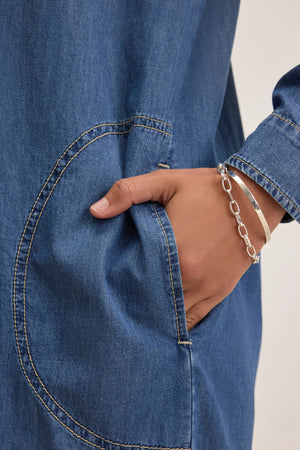 A person in a Velvet by Graham & Spencer IZMIR CHAMBRAY DRESS, with a hand in the pocket, wearing a silver chain bracelet.
