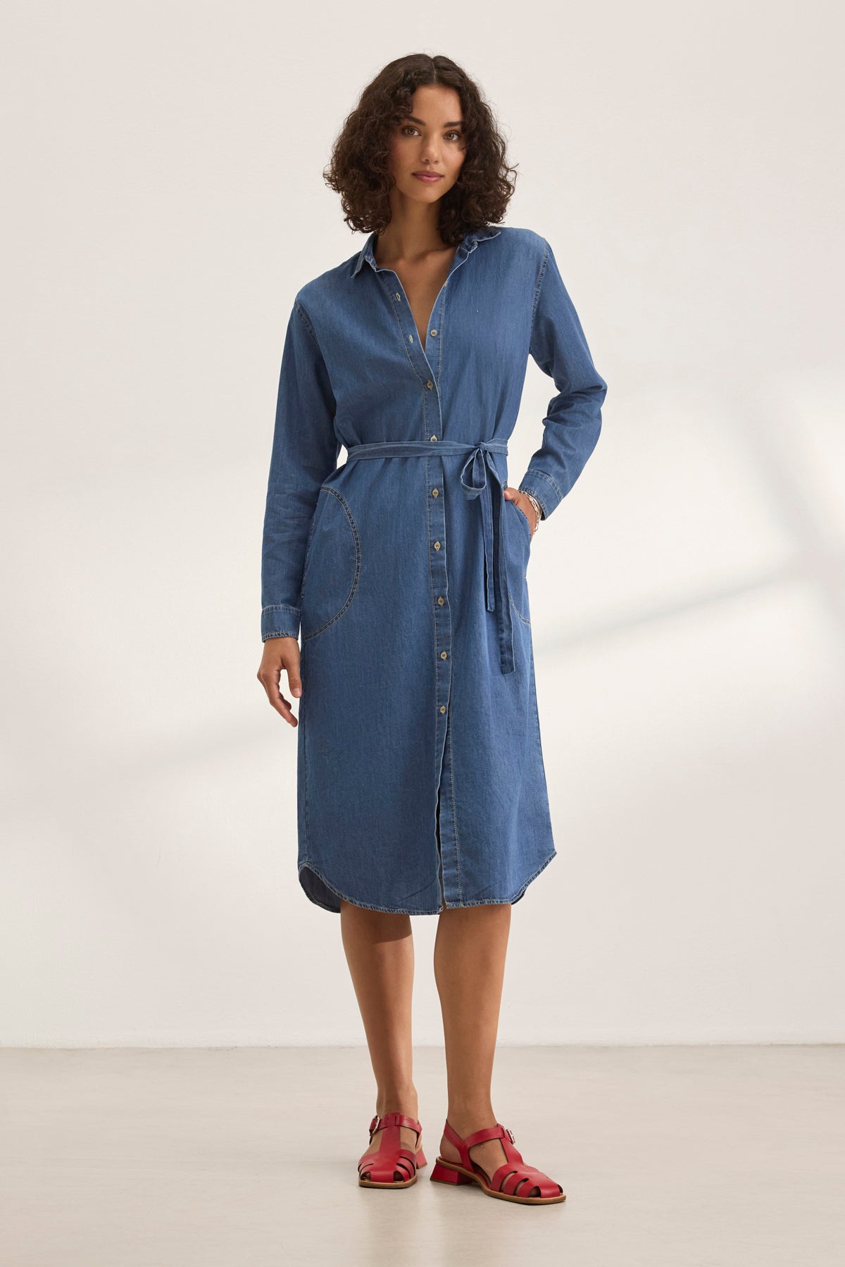 A woman in the IZMIR CHAMBRAY DRESS by Velvet by Graham & Spencer, paired with red sandals, stands against a plain white background.-38820646355137