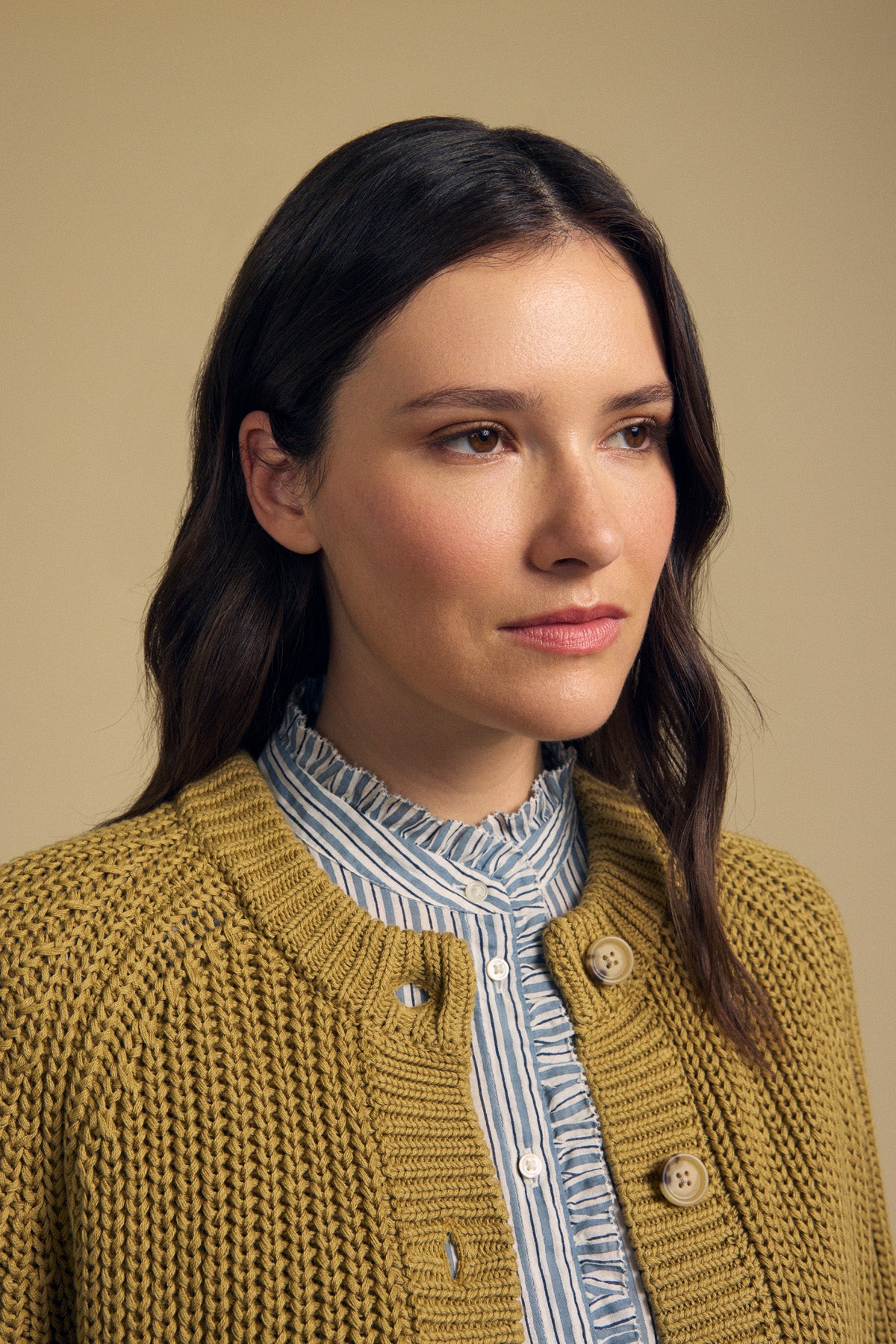 A woman with long brown hair models the Velvet by Graham & Spencer LISABETH CARDIGAN, a chunky cropped style layered over a blue striped shirt, standing against a plain beige background.-41940441170113