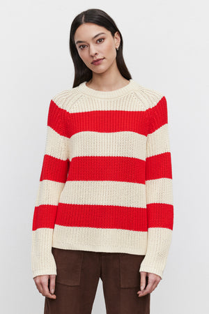 A person wearing the CIARA SWEATER from Velvet by Graham & Spencer, featuring cream and red horizontal stripes in a cotton knit fabric, is paired with brown pants and stands against a plain white background.