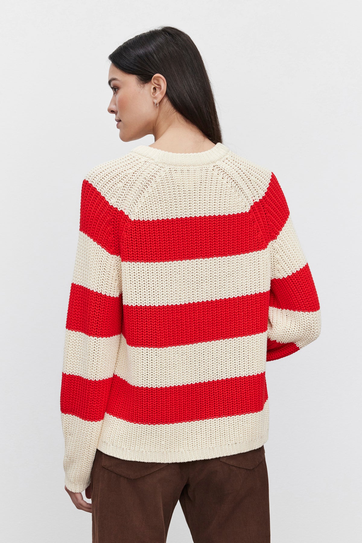 A person with long hair stands facing away, wearing the CIARA SWEATER from Velvet by Graham & Spencer—a red and white cotton knit sweater with a striped design—and brown pants against a plain background.