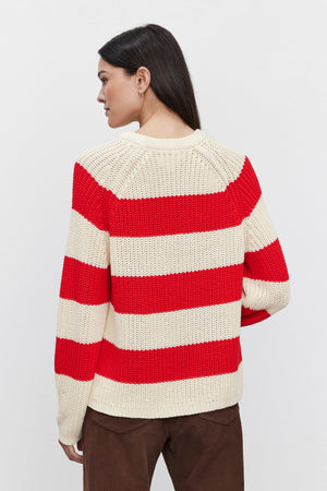 A person with long hair stands facing away, wearing the CIARA SWEATER from Velvet by Graham & Spencer—a red and white cotton knit sweater with a striped design—and brown pants against a plain background.