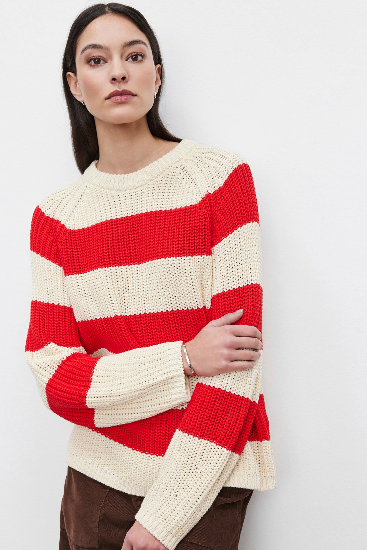 A person with long dark hair is wearing the CIARA SWEATER by Velvet by Graham & Spencer, featuring a cream and red cotton knit with a striking striped design, while posing against a plain white background.-37665553678529
